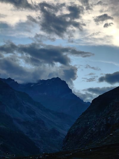 An upward angle shot captures a dramatic mountain landscape under a cloudy sky. The image is dominated by dark, imposing mountain ranges, their peaks partially obscured by shadow and mist. The sky above is a mix of deep blues and grays, with lighter patches indicating breaks in the cloud cover. In the foreground, the slopes of the mountains are steep and rugged. The lower part of the image shows a dimly lit valley floor with a few scattered buildings and vehicles, suggesting a small settlement or roadside area. The overall mood is one of rugged natural beauty and perhaps a sense of remoteness. The location is identified as Damphug, India.