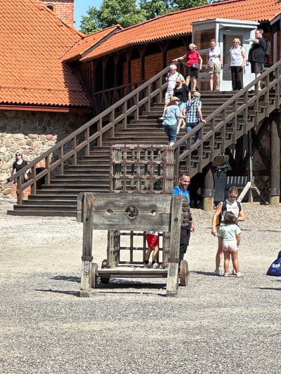 The image depicts an outdoor scene at Trakai Island Castle in Trakai, Lithuania. In the foreground, a wooden pillory, an archaic punishment device, stands on a gravel-covered ground. Behind it, a wooden scaffold structure with metal bars is visible. A man in a blue shirt and black pants stands to the right of the pillory, looking to his left. A woman in a white shirt and shorts with a backpack stands next to a young girl in a light green shirt and pink shorts. Both are looking towards the man. In the background, a wooden staircase with railings leads up to a building with a red tiled roof. Several tourists are visible on the stairs, some ascending and some descending. The environment appears to be a sunny day, with shadows cast on the ground.