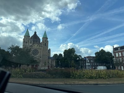 The image depicts a street view in the city of Maastricht, Netherlands. In the foreground, a section of a road is visible with a white line marking the lane. Behind the road, there is a strip of vegetation with green and yellow foliage, some dry grass, and flowering plants.

Dominating the left side of the mid-ground is a large stone church with a circular stained-glass window above its entrance. The church has two prominent green-spired towers on either side of a darker, domed central section, topped with a cross. Trees partially obscure the church and the surrounding buildings.

In the background, several buildings are visible, including a multi-story building with arched windows and a red-tiled roof, and a brick building with white window frames and dark roofing. A white van with the logo "apleco" is parked in front of one of these buildings. The sky is blue with scattered white clouds and contrails from aircraft. The overall impression is a sunny day in an urban environment with a prominent religious structure.