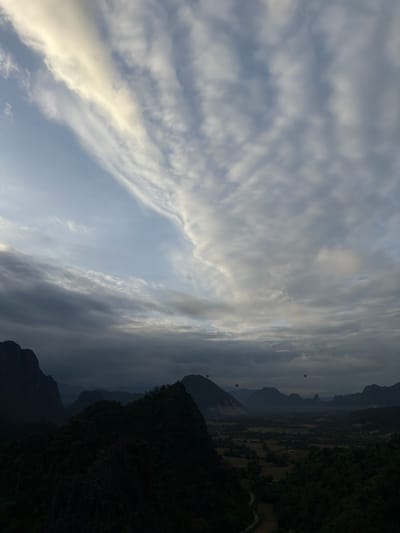 The image depicts a panoramic view of a mountainous landscape at dawn or dusk. In the foreground, dark, densely forested mountains dominate the lower half of the frame. The terrain is rugged with visible rock formations. As the eye moves towards the background, the landscape transitions into a valley dotted with cultivated fields, suggesting an agricultural area. Several hot air balloons are visible in the sky, dispersed across the middle ground, indicating recreational activity. The sky itself is a dramatic display of clouds, with streaks of lighter clouds suggesting the sun's rays breaking through or the lingering light of twilight. The overall atmosphere is serene and majestic, capturing the natural beauty of the location. The scene is likely from the outskirts of Ban Phon Sai, Laos, a region known for its karst mountains and hot air ballooning activities.