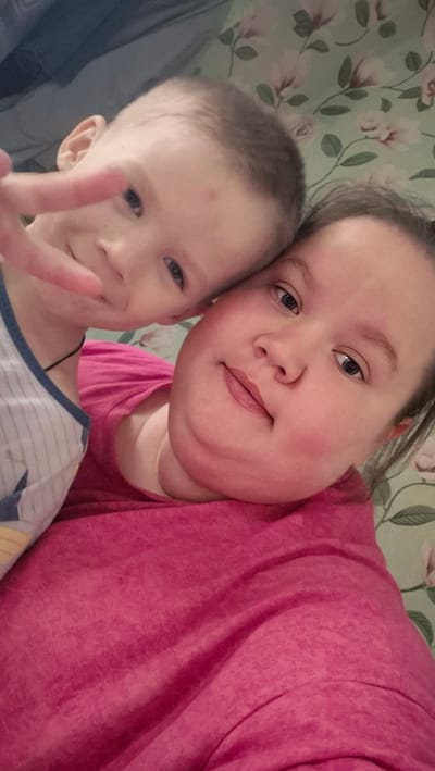 A selfie-style photograph shows two individuals: a child giving a peace sign and an adult. The child, positioned on the left side of the frame, has short light brown hair and blue eyes. The child is wearing a white and blue striped shirt. The adult, to the right of the child and slightly behind, has light brown hair and blue eyes. The adult is wearing a bright pink top. The background features a floral patterned wallpaper with light green and white tones. The location is Большие Кусты, Russia.
