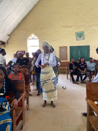 This media file depicts a gathering of people, predominantly women, inside a room that appears to be a church or a community hall. The subjects are seated on wooden benches, some are standing. In the foreground, a woman in traditional attire, including a headscarf and a patterned skirt, is prominently featured. She wears a necklace of colorful beads. Behind her, other individuals are visible, some in casual clothing, others in more traditional outfits. The walls of the room are a pale yellow, and there is a green door and a window with natural light entering the space. A crest with a cross and the words "HOSPITAL VOM" and "N C" is visible on the wall, along with a small plant placed on the floor. Several instances of text are present in the image, including "VICTORY" on a shirt, and "PDA GICHI BECHEH ZADSWORD" on a cloth, and "EVEL VE IRIGW T ASSOCIAT" on a blue patterned fabric. The overall atmosphere suggests a religious service, a celebration, or a community meeting. The time of day is not explicitly indicated, but the natural light suggests daytime. The location cues, such as the crest and the style of the building, point to Dutsen Kato, Nigeria.