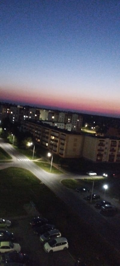 The image is an aerial view of a residential area in Skidzieĺ, Belarus, at dusk. The sky displays a gradient of blue and pink hues, transitioning from a light blue at the top to a pinkish-purple band near the horizon. Below the sky, a row of multi-story apartment buildings stretches across the frame, their windows illuminated with warm light, indicating occupancy. The buildings appear to be of Soviet-era architecture.
In the foreground and middle ground, streets and intersections are visible, marked with streetlights casting pools of light on the asphalt. A crosswalk is clearly discernible on one of the roads. Several parked cars are scattered throughout the scene, some on the street and others in parking lots adjacent to the buildings. These vehicles include sedans and vans of various colors.
Green grassy areas are interspersed between the buildings and the roadways, suggesting a planned urban landscape. The overall impression is of a quiet, ordinary evening in a suburban neighborhood of Skidzieĺ. No people are clearly visible in the image, though a dark figure, possibly a person, is faintly discernible in the lower right corner. The lighting conditions suggest it is either early morning or late evening.