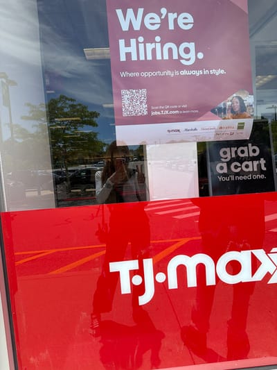 The image shows a reflection on the window of a TJ Maxx store, indicating the location is in Orleans, United States of America. A woman is taking a selfie, holding her phone and making a peace sign with her hand. She is wearing a white long-sleeved shirt, dark pants, and sneakers. Reflected in the window are trees, sky, and a parking lot with cars. A large "We're Hiring" sign is visible, along with a QR code and the website jobs.TJX.com. Below that, the TJX family of brands including TJ Maxx, Marshalls, HomeGoods, and Sierra are listed. Another sign reads "grab a cart. You'll need one." The lower portion of the image features the red TJ Maxx logo.