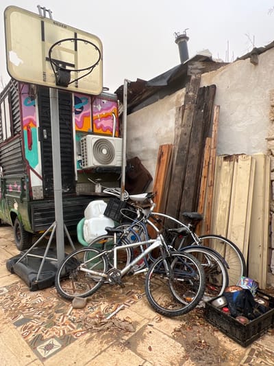 This image depicts an outdoor area in Jisr az-Zarqa, Israel, characterized by a dense arrangement of objects against a building wall under an overcast sky.
In the left foreground, a portable basketball hoop with a translucent rectangular "SILVER" and "SPALDING" branded backboard and a black metal hoop stands on a large black plastic weighted base. To its right, a cluster of at least three bicycles is present; a silver mountain bike with black tires and disc brakes is most prominent, leaning against a darker blue-grey bicycle.
Adjacent to the basketball hoop and bicycles, a customized olive-green utility truck or van is parked. Its rear section features corrugated metal paneling and a side adorned with vibrant, abstract graffiti-style artwork in pink, orange, yellow, teal, and blue, including planet motifs. A white outdoor air conditioning unit is mounted on the truck's side. Several white plastic containers and a black plastic crate are stacked near the truck's base.
The ground surface is paved with weathered ceramic or concrete tiles featuring geometric patterns in tones of brown, beige, white, and blue-grey. Scattered leaves and dirt are visible across the tiled surface. In the lower right foreground, a black plastic crate contains miscellaneous items, including dark fabric and a metal pot.
The background consists of a light-colored, textured building wall, possibly stucco or plaster, against which numerous pieces of lumber and wood planks of varying sizes and conditions are stacked vertically. Above the wall, a makeshift roof structure of corrugated metal is visible, from which a dark-colored chimney or vent pipe extends upwards. The sky above is uniformly light grey and overcast. No individuals are present in the scene.