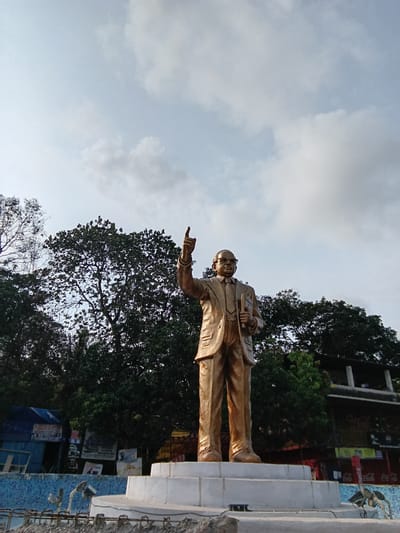 A low-angle shot captures a bronze statue of a man in a suit, likely Dr. B.R. Ambedkar, atop a white pedestal in Chopdem, India. The man wears glasses and holds a book in one hand, while pointing forward with the other. Behind the statue, dark green foliage fills the frame. The cloudy sky is visible in the upper portion of the shot. In the background, there are buildings with signage and advertisements. The base of the statue and the surrounding area appear to be under construction, as indicated by exposed rebar and unfinished mosaic work.