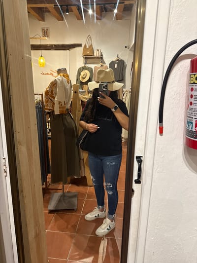A reflection in a mirror shows a person taking a picture of themselves in a clothing store in San Cristóbal, Mexico. The person is wearing a black t-shirt, jeans with rips, and white sneakers. They are wearing a straw hat and an Apple Watch. The store has clothing racks and various articles of clothing, including tops, pants, and hats. A fire extinguisher is mounted above the mirror frame. The floor is tiled. A single pendant light fixture hangs in the store.