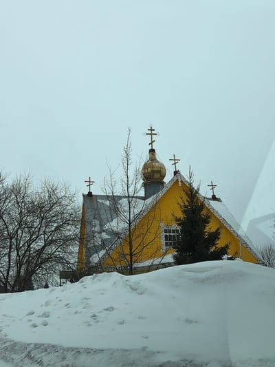 A vibrant yellow, wooden church stands partially obscured by a tall snowdrift. The building features ornate trim and multiple Orthodox crosses atop its spires. Bare trees frame the structure, adding a stark contrast to the bright color. A painted banner hangs on the side of the church. The background is a muted sky, indicative of winter.