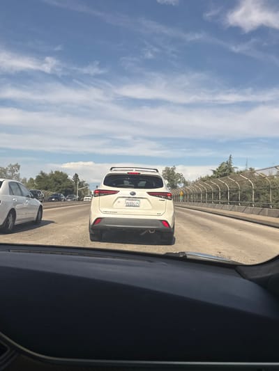 The image shows a view from inside a vehicle on a highway, likely in Citrus Heights, California, as indicated by the license plate frame on the white SUV. The primary focus is the rear of a white Toyota Highlander hybrid SUV with a roof rack, driving in the middle lane of a multi-lane highway. The license plate reads "8VBB415". To the left of the SUV, a white sedan is visible in the left lane. Further ahead, several other vehicles are present on the highway. The road is paved, with clear lane markings. On the right side of the highway, there is a concrete barrier with a chain-link fence on top, separating the road from vegetation and trees. The sky is blue with scattered white clouds. The overall scene depicts a typical traffic situation on a sunny day.