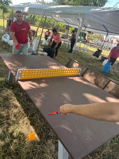 The image depicts an outdoor gathering under a white canopy, likely in Lovets, Bulgaria. A man in a red t-shirt and glasses is holding a red table tennis paddle, appearing to be playing. Nearby, a woman in a pink top and black pants is also present. A table tennis table with a yellow net is set up, and an orange ping pong ball rests on the grass in the foreground. Several other people are visible in the background, along with various objects including chairs, bags, buckets, and a blue gas cylinder. The environment is grassy with some trees and sunlight filtering through.