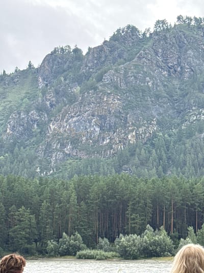 The image shows a view of a large, rugged mountain covered in green vegetation and trees. The sky above is overcast and gray. In the foreground, there is a body of water, likely a river or lake, with ripples on the surface. On the left side of the foreground, the back of a person's head with brown hair is visible. On the right side of the foreground, the back of a person's head with blonde hair is visible. The overall scene suggests an outdoor, natural environment in Nizhnekayancha, Russia.