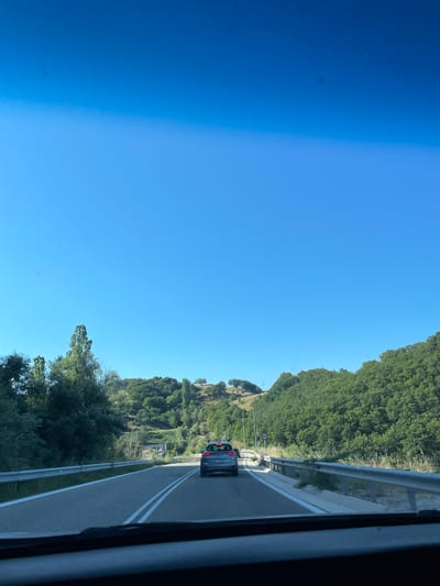 The image shows a view from inside a vehicle driving on a road. The road is paved and has two lanes separated by white lines. On the right side of the road, there is a guardrail and a roadside ditch. The area is hilly and covered with lush green trees and vegetation. The sky is clear and blue. In the distance, a silver car is driving ahead on the same road. A speed limit sign indicating "60" is visible on the right side of the road. The overall scene suggests a drive through a rural or semi-rural landscape. No specific landmarks or details are visible to indicate the location is Komotini, Greece.