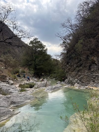 The image displays a valley scene in San Miguel Atlapulco, Mexico, under an overcast sky with scattered grey and white clouds. A natural waterway, characterized by multiple pools of clear, light turquoise-green water, flows through the center foreground and middle ground, bounded by large, smooth, grey-white rock formations.
Steep, rocky hillsides flank both sides of the valley. The hillsides are covered with a mix of vegetation, including numerous deciduous trees with bare branches, particularly on the upper slopes, and denser evergreen foliage closer to the valley floor. Sparse shrubs and grasses grow among the rocks and along the water's edge. On the right side, a dark, rocky cliff face rises steeply, displaying some small agave-like plants.
Approximately 10 to 15 individuals are present within the scene. On the left bank, in the middle distance, a dirt path leads towards a rudimentary shelter with a yellowish-tan tarp roof. A group of approximately four individuals are walking on this path near the shelter. Other individuals are dispersed along the rocky banks of the waterway, standing or sitting. One individual in dark clothing is visible near the water's edge against the rocky cliff on the far right. A thin, light-colored rope or cable stretches horizontally across the upper middle ground, attached to the right cliff face.