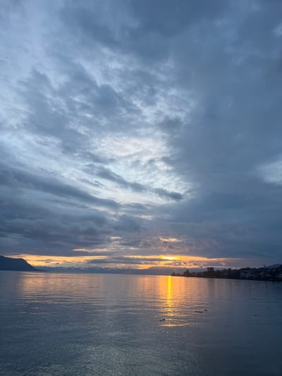 The image depicts a large body of water, identified as Lake Geneva in Montreux, Switzerland, under an overcast sky during sunset. The sky is dominated by dark grey and blue stratified clouds, with a distinct horizontal band of yellow and orange light on the horizon where the sun is setting. This solar light creates a prominent, elongated reflection of orange and yellow on the water's surface, extending from the horizon towards the right foreground. Mountains are visible on the far left horizon, while the urban coastline of Montreux, featuring buildings and structures, extends along the right horizon. Multiple small, dark, indistinct objects are observed floating on the water in the right foreground. No human figures, animals, or active subjects are present within the frame.