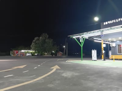 The image depicts an outdoor scene at night, likely a service area or a small complex, in the city of Sayat Nova, Armenia. A prominent feature is a modern, covered structure with bright, strip lighting along its underside, supported by vibrant green poles. The structure appears to house some form of service equipment, as evidenced by white charging stations or similar utility units. A yellow bench is visible to the right of the structure. The ground in the foreground is asphalt, marked with white dashed lines and yellow directional arrows indicating traffic flow. In the background, trees and a small, red, shed-like building are visible. A white and a dark-colored car are parked in the distance, near a colorful advertisement or display. The sky is dark, with a bright light source creating a lens flare effect in the upper right portion of the image, possibly a streetlamp or a light from the structure. The overall impression is one of a well-lit, functional outdoor space at night.