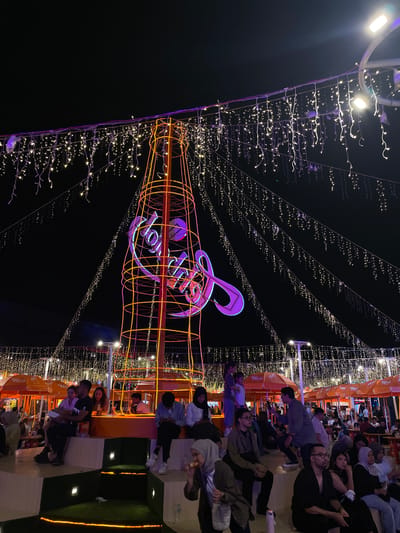 The image shows an outdoor scene in Salembaran Jati, Indonesia, at night. A large, illuminated structure resembling a Coca-Cola bottle with a musical note and the word "Toba Dream" stands prominently in the center. The bottle is constructed with orange neon lights. Decorative string lights hang overhead, creating a festive atmosphere. A crowd of people is seated on stepped seating areas on the right side of the image, possibly watching or enjoying the illuminated display. Orange umbrellas are partially visible above the seating areas. The background is dark, emphasizing the lighted structure and surrounding area.