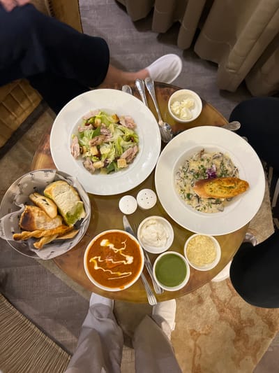 An overhead photograph details a dining scene within a Radisson hotel restaurant or lounge, located in Noida, India. The primary subject is a round, reflective golden-hued table laden with various dishes and accompaniments for multiple diners.
Three individuals are partially visible: one on the left with dark trousers, one on the far right in dark trousers, and one at the bottom center wearing light-colored trousers and white slippers.
The table holds:
- A white plate on the upper left featuring a Caesar salad with lettuce, croutons, and flaked chicken or tuna.
- A white plate on the upper right containing a creamy mushroom dish, likely risotto or pasta, garnished with herbs and a slice of toasted bread.
- A white bowl on the lower left with a reddish-orange curry or soup, embellished with white cream drizzles.
- A metal basket lined with paper, clearly branded "Radisson," holding assorted breads including focaccia-style bread and breadsticks.
Additional items include:
- A small white bowl with pats of butter positioned on the upper right.
- White salt and pepper shakers situated centrally.
- Three small white bowls on the lower right: one containing a white creamy dip, one a green chutney, and one a yellow granular substance (possibly grated cheese or roasted lentil crunch).
- Silver cutlery, including ornate spoons, forks, and a knife, is arranged around the plates.
The environment features carpeted and hard flooring, with upholstered seating visible on the left and upper right, consistent with a hotel dining area.