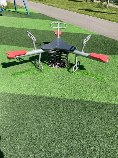 An overhead shot captures a spring-loaded seesaw on a playground. The seesaw has two red seats, each with a heart-shaped cutout, and a grey saddle-like seat in the center. The seesaw is supported by a metal frame and heavy springs. It is situated on a green, artificial turf surface, with strips of darker green artificial turf and a grey paved path visible in the background. The scene appears to be in a park or recreational area in the city of Zakharino, Russia.