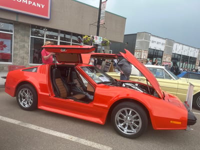 This media file depicts an outdoor car show, likely held in the city of Leduc, Canada, given the visible text. The primary focus is a bright orange sports car with distinctive gull-wing doors, positioned prominently in the foreground. The car's hood and doors are open, revealing its interior and engine components, suggesting it's on display.
Several people are present, casually observing the vehicles. One man, wearing a straw hat and a red t-shirt, is seen near the open door of the orange car. Behind it, a yellow classic car is also visible, indicating multiple vehicles are part of the event. The background shows a street with buildings, including one with a red sign displaying the word "COMPANY" in large white letters. Another building appears to be under construction, with visible framing and sheathing.
Street signs and lampposts are present, with one lamppost holding hanging flower baskets and parking signs. A banner on the lamppost reads "MAIN STREET" and "Leduc," confirming the location. Another sign reads "TAPROOM OPEN." The weather appears overcast, suggesting a cloudy day. The overall atmosphere seems to be that of a community event, likely a car enthusiast gathering or a local festival. The time of day is difficult to ascertain precisely but appears to be daytime.