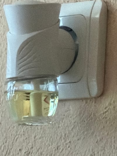 An illuminated, close-up image taken in Breznitsa, Bulgaria, depicts a white plug-in air freshener device inserted into a white electrical wall outlet. The air freshener's upper portion is composed of opaque white plastic, featuring a curved, ribbed design on its frontal surface. Attached beneath is a transparent glass or plastic reservoir, approximately half-filled with a translucent yellow liquid. A white wick is visible extending upwards from within the liquid into the device's housing. Reflections, including a partial red "Z" symbol, are discernible on the liquid's surface and through the clear container. The electrical outlet is white, exhibiting two round socket apertures, with one occupied by the air freshener. The outlet is affixed to a wall characterized by a textured, light beige, or off-white finish. No individuals, actions, or interactions are depicted.