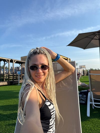A woman with blonde braided hair, wearing sunglasses and a black and white patterned swimsuit, is posing for a selfie while sitting in a reclining chair outdoors. She is holding her hair with one hand and her left arm is resting on the chair. She is wearing a blue wristband on her left wrist. The background shows a grassy area with lounge chairs, umbrellas, and some wooden structures. The sky is blue with a few wispy clouds. The location is implied to be Dumino, Russia, although specific landmarks to confirm this are not present in the image.