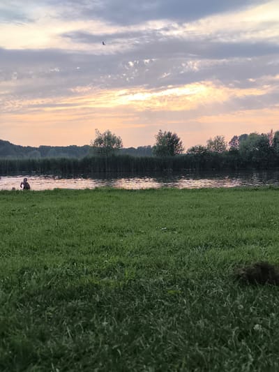 The image depicts a tranquil scene by a body of water, likely a canal or small lake, at dusk in Broek op Langedijk, Netherlands. A person, possibly a child given their size and apparent activity, is partially submerged in the water on the left side of the frame, with another spherical object near them in the water. The water's surface reflects the soft, pastel colors of the sunset sky, which is characterized by scattered clouds tinged with pink and orange. A line of dense greenery, including reeds and trees, forms the far bank of the water. In the foreground, a field of lush green grass stretches out, with a small pile of dark material, possibly soil or debris, visible on the right. The overall atmosphere is peaceful and natural.