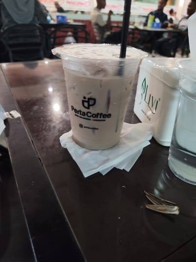 A medium shot captures an iced coffee drink in a clear plastic cup placed on a dark, glossy table. The cup, branded with a black logo that reads "PertaCoffee" and includes a stylized 'P', is topped with a clear plastic lid and a black straw. Condensation droplets are visible on the exterior of the cup. The drink appears to be a light brown, milky color. Beneath the cup is a white paper napkin. To the right of the coffee cup is a white plastic container with a green logo that says "Livi" and a website address below it. Further to the right, a glass of water is partially visible. In the background, the scene is blurred, but suggests a cafe or restaurant setting with other patrons and tables. The overall lighting is somewhat dim, with highlights reflecting off the table surface. The location is specified as Meunasahceubrek, Indonesia.