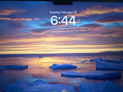 A screen displays Tuesday, February 10 at 6:44. Below the time and date, a vibrant background depicts a body of water dotted with icebergs. The sky transitions from a yellow horizon line to blues and purples with streaks of white clouds. The water reflects the colors of the sky, creating a shimmery surface where the icebergs sit. The icebergs are various shapes and sizes, appearing primarily in shades of blue.