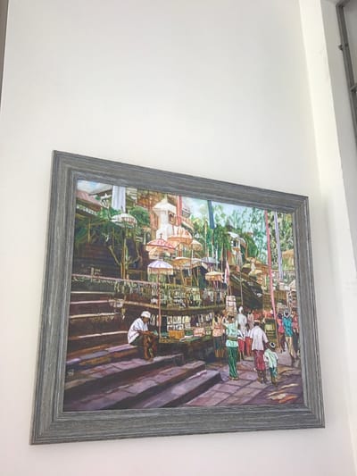 This image is a photograph of a framed painting mounted on a plain, light-colored wall, taken in the city of Kota Batu, Indonesia.
The painting depicts a bustling outdoor scene, characteristic of a Balinese temple complex or market. In the lower left and extending upwards, a series of wide, dark-toned stone steps forms a prominent feature. On these steps, towards the left, a single man is seated, wearing a white head covering and a brown sarong. To the right of the steps, a crowded pathway is visible, filled with numerous individuals. Many are dressed in traditional attire, with several figures carrying items balanced on their heads, consistent with either market goods or ceremonial offerings. Market stalls, displaying various items, line this pathway.
The mid-ground and background are densely populated with numerous tall, ornate decorative umbrellas (Tedung), predominantly in shades of white, orange, and red, featuring fringed edges. These are typically associated with ceremonial or sacred sites. Behind these umbrellas, traditional stone temple architecture, including what appear to be gate-like structures and shrines, is visible, interspersed with lush green foliage. Vertical, multi-colored banners or flags are also present, contributing to the vibrant atmosphere.
The painting is enclosed within a rustic, grey-brown, wood-textured frame.