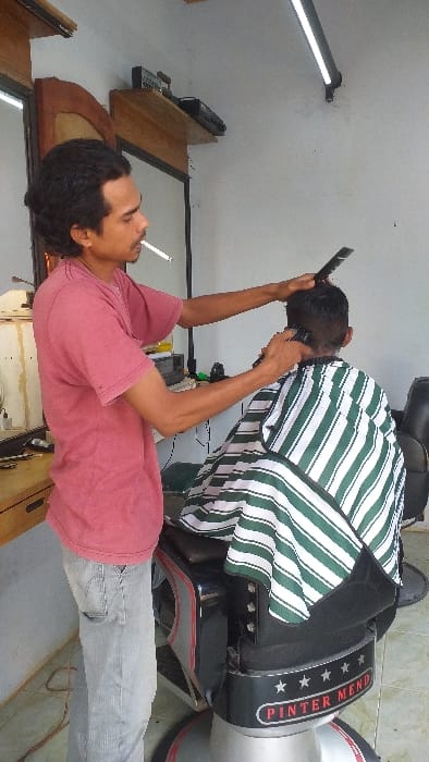 Within a barber shop in Serdang, Indonesia, a male barber is cutting a male client's hair. The barber, with a lit cigarette in his mouth, wears a reddish-pink t-shirt and light grey jeans, using an electric hair clipper in his right hand and a black comb in his left. The client, viewed from behind, is seated in a black and silver barber chair, draped in a white and dark green striped cape. The barber chair's base features "PINTER MEND" and five stars. The scene includes a white wall background, a wooden-framed mirror, a wooden shelf holding electronic equipment, and a horizontal LED light fixture mounted on the upper wall. A brown electrical cord is coiled on the tiled floor.