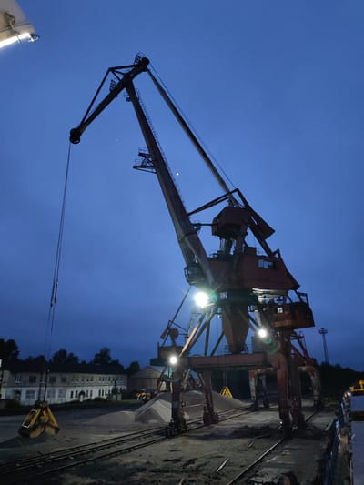 This image depicts an industrial port or yard at dusk or night, characterized by a deep blue sky and illuminated by artificial lights. A large, rusty gantry crane dominates the scene, its intricate structure and control cabins highlighted by several bright floodlights. The number "52" is visible on the side of this central crane.

To the left, another crane's boom extends, suspending a large, yellow clam-shell grab bucket poised above a substantial pile of light-colored aggregate material, likely sand or gravel. Multiple similar piles are scattered across the ground. A network of railway tracks runs across the foreground and mid-ground, indicating a transport hub for bulk goods. In the distance on the left, a long, multi-story industrial building with numerous windows appears weathered. A yellow loader vehicle can be seen in the background near the material piles, and a slender tower stands against the sky on the far right. The lower right corner reveals part of a ship or dockside structure.

The clear, dark blue sky features a faint crescent moon or bright star visible near the top of the main crane's structure. The artificial lighting creates an operational atmosphere, illuminating the heavy machinery and the industrial environment. No people are explicitly visible. The scene suggests active loading or unloading operations of raw materials in this industrial setting, likely a port facility in Medvezhyegorsk, Russia.
