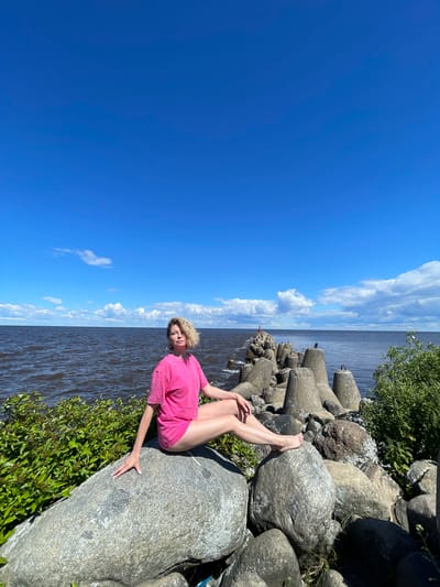 A woman with blonde hair and a pink shirt sits on large rocks by a body of water under a bright blue sky with scattered clouds. She is positioned on a large grey boulder, with her legs extended. Her right leg is bent at the knee, and her left leg is extended towards the water. She is looking towards the camera with a relaxed expression. The background shows a long, concrete breakwater stretching into the water, and dense green foliage. The water is dark and appears slightly choppy, reflecting the bright sky. The scene suggests a warm, sunny day, likely in the late morning or early afternoon. The presence of the breakwater and the type of vegetation hint at a coastal or large lake environment. The overall mood is peaceful and serene. There is no visible text in the image.