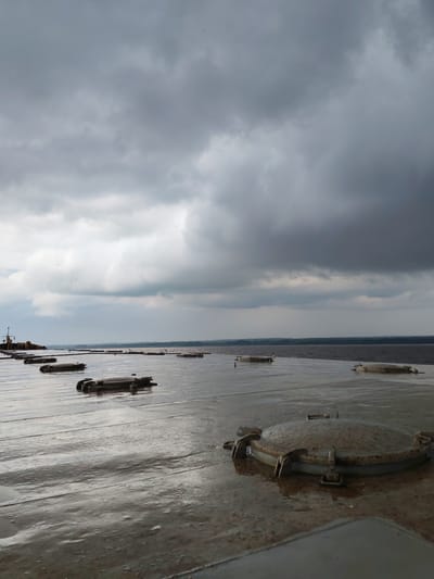 The image shows a vast, overcast sky above a flat, wet surface with several circular, metal hatches embedded within it. The surface appears to be the deck of a ship or barge, with reflections of the sky and hatches visible on its wet, metallic sheen. In the distance, a body of water stretches to the horizon, bordered by a dark line of trees or land. On the far left, a portion of a structure, possibly another vessel or a dock, is visible. The overall atmosphere is somber and industrial, likely captured during or after rain. The specific location in Shchetinskoe, Russia, is not visually identifiable from the image alone, but the scene suggests a watery, possibly industrial or transportation-related environment.