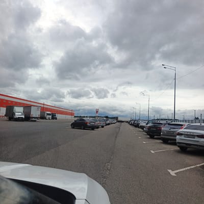 An overcast day in a parking lot outside a large building complex in Sof'ino, Russia. The building has a long, orange and white facade with many windows and is likely a warehouse or retail establishment. Several white trucks are parked along the building. The parking lot is paved with asphalt and marked with white parking lines. A row of cars is parked in the lot, facing away from the building. Some of the cars appear to be Russian-made Lada models. Streetlights are visible in the distance. A white car, possibly a sedan, is partially visible in the foreground, suggesting the image was taken from inside a vehicle. The sky is heavily clouded with grey and white tones.