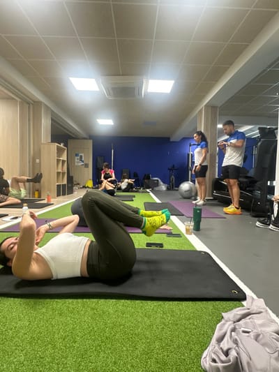 Inside a well-lit gym, several people are exercising. In the foreground, a woman lies on a black mat with her legs raised, wearing green leggings and patterned socks. Behind her, others are on mats, while farther back, individuals use treadmills and exercise bikes against a blue wall. Lockers and storage shelves line one side of the room. The gym floor alternates between gray, purple yoga mats, and artificial turf.