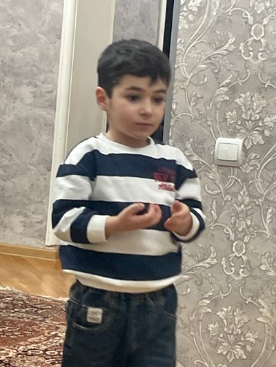 A boy stands indoors. He is wearing a striped shirt and jeans. The walls are patterned with floral designs. A light switch is on the wall behind him. Part of a rug is visible on the floor. No location data is available.