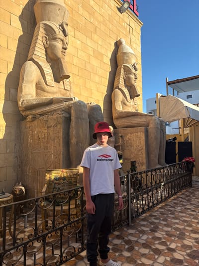 A medium shot presents a young male standing in front of an Egyptian-themed building. He is wearing a white t-shirt with "Balenciaga" printed on it, dark pants, a red bucket hat, and white Crocs. He has a pink wristband on his left wrist. He is standing on a staircase with a decorative metal railing. Behind him are two large, tan-colored sarcophagi, intricately carved with Egyptian hieroglyphs. The building itself is a tan color, constructed of stone. The sky is clear and blue. A partial view of the building above the sarcophagi can be seen, consisting of white, slanted sections and a partial view of a black awning.
Based on the architectural style and the sarcophagi, the location is likely Dahab, Egypt, as it is a popular tourist destination known for its Egyptian-themed establishments.