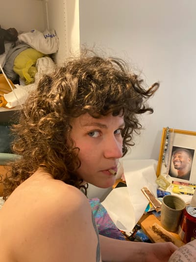 The image shows a person with curly brown hair and light skin looking at the camera. The person has something in their mouth, possibly a cigarette or joint. The background appears to be a cluttered space. On a surface behind the person, there is a framed picture of a man, a ceramic mug, a can, rolling tobacco, rolling papers, and what appears to be a box of condoms. To the left of the person, there is a closet with clothing items visible inside. The location of Le Pré-Saint-Gervais, France is not inferable from the image.