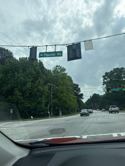 The image shows a street view, likely taken from inside a vehicle. A green street sign indicates "Pounds Rd" with a left arrow. Traffic lights are visible, with one set showing green. Several vehicles are on the road, including a white truck and a dark-colored car. The sky is overcast. The location is identifiable by the street sign as the city of Mountain Park, United States of America.