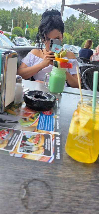 A woman with dark, up-styled hair is captured from the chest up, holding a smartphone in her right hand, seemingly taking a photo or video. She is wearing an off-the-shoulder white top. Her focus appears to be on the phone, which displays a small blue and white card with the text "Miss Pepper" and a cartoon image of a woman. The phone is positioned in front of a drink served in a mason jar, which is topped with slices of watermelon and orange, and a small green fruit.
On the table in front of her, to her left, is a black ashtray containing cigarette butts. To the left of the ashtray, a salt and pepper shaker set is visible. The table surface is a wooden veneer with a visible water ring.
In the background, out of focus, are cars and trees, suggesting an outdoor dining setting. The sky is overcast with clouds. A menu or placemat with food images and text in German is laid out on the table. To the right of the frame, a second mason jar drink, this one yellow and containing fruit and a straw, is partially visible. Based on the context of the menu and the "Miss Pepper" branding on the phone, this scene likely takes place at a restaurant or cafe in Wietzendorf, Germany, possibly one associated with the "Miss Pepper" establishment.