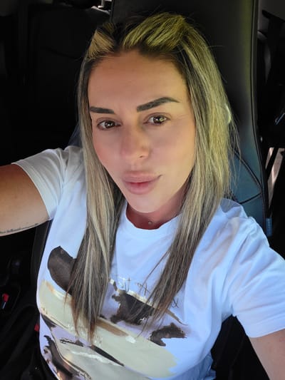 An outdoor, eye-level, medium shot captures a fair-skinned woman with long, blonde and brown highlighted hair, styled down her shoulders. She is looking at the camera with a neutral expression. She is wearing a white t-shirt with a abstract, gold, black, and white design on it, a silver necklace, and a single silver piercing below her lip. The woman is sitting in the driver's seat of a car, suggested by the black leather seat and the seatbelt buckle visible on the left side of the frame. The interior of the car is dark, with no other occupants visible. The lighting suggests it is daytime. The background is dark and indistinct. There are no landmarks or identifying features to indicate a specific location within Carpi, Italy, or anywhere else.