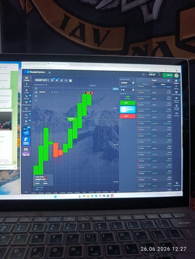 The image shows a close-up of a laptop screen displaying the PocketOption trading platform. The screen shows a candlestick chart of EUR/JPY OTC currency pairs, with the current time displayed as 12:27:12 UTC+3. The chart indicates a trading session with green and red candlesticks, suggesting buying and selling activity. The right side of the screen lists various trade executions with timestamps, asset names, amounts, and profit/loss figures.
The laptop is placed on a surface that appears to be a desk, and a portion of a keyboard is visible in the foreground. The overall scene is dimly lit, with the glow of the laptop screen being the primary light source. The background of the laptop screen features a scenic image, possibly a mountain landscape.
Visible text includes the trading platform's name "PocketOption", currency pair "EUR/JPY OTC", various trade details such as "Tesla OTC +92%", "Apple OTC +47%", and specific monetary values in UAH. There are also timestamps and navigation labels like "Trading", "Finance", "Profile", "Market", "Buy", and "Sell". A date stamp "26.06.2026 12:27" is visible on the lower right corner of the image.
The context provided indicates the location as Novovolynsk, Ukraine. The image captures a moment of active online trading, with the user presumably monitoring financial markets.