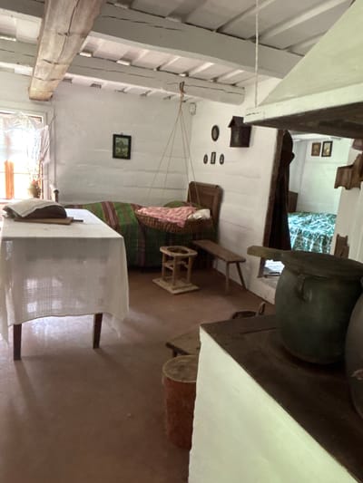 This is a photograph of the interior of a historic dwelling in Sielachowskie, Poland. The room is sparsely furnished, suggesting a rustic and traditional setting. Visible elements include a white tablecloth draped over a table in the foreground, a wooden bed with a green and red striped blanket, and a suspended crib. On the walls, there are small framed pictures and what appear to be decorative objects or possibly old light fixtures. A large, dark pot sits on a hearth in the right portion of the frame. The walls are white and appear to be constructed of logs or rough plaster, and wooden beams are visible on the ceiling. The lighting suggests it is daytime, with light filtering in from a window on the left. The overall impression is of a preserved historical domestic space.