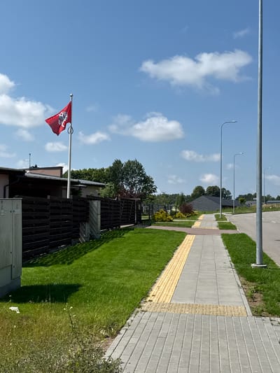 The image depicts an outdoor scene on a clear, sunny day with scattered white clouds. A red flag with a white emblem is flying from a flagpole, which stands near a building with a dark wooden fence. To the right of the fence is a well-maintained green lawn. Adjacent to the lawn is a paved walkway with textured yellow tiles, suggesting a pedestrian path or tactile paving. Beyond the walkway, a road is visible, with streetlights lining its side. The background features trees and more distant structures. The overall setting appears to be a suburban or residential area in Mažoji Riešė, Lithuania. No people are visible in the frame. The time of day is likely daytime, given the bright sunlight casting shadows. The weather is pleasant and sunny.
