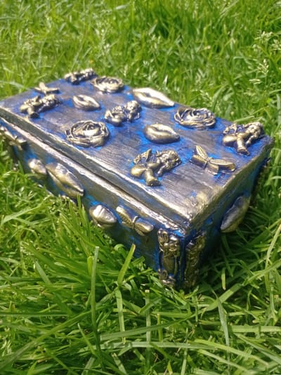 A decorated blue and gold rectangular box rests on a patch of green grass. The box is adorned with various raised embellishments, including what appear to be gold roses, lips, dragonflies, and cherub-like figures. The box is positioned at an angle, with its lid slightly ajar. The background is a blur of more green grass, indicating an outdoor setting. There is no indication of a specific location within Дукорскі сельскі Савет, Belarus.
