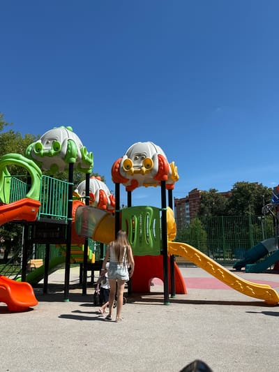 An outdoor playground is depicted on a sunny day with a clear blue sky. The playground features a large, multi-colored structure with slides and climbing elements. The structure is primarily red, yellow, and green, with whimsical, grey and orange, creature-like decorations at the top. In the foreground, a young person with long, blonde hair, wearing a white tank top and denim shorts, stands facing away from the camera, looking at the playground. To their left, a stroller is visible with a child in it. The ground surface of the playground is paved with a light-colored, textured material, with circular red and grey markings on it. In the background, there is a green metal fence, trees, and apartment buildings. The location is identified as Щапово, Russia.