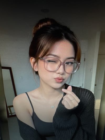 A young woman with her hair pulled up in a bun, wearing glasses and a dark sweater over a grey camisole, is shown from the chest up. She is winking and making a heart shape with her fingers. A silver necklace with a butterfly pendant is visible. A mirror with a wooden frame is in the background, to the left of the woman. The environment appears to be indoors, with a plain wall behind her. The image is a screenshot, as indicated by the pixelated appearance and faint reflections on the screen. The location of Protasovo, Russia is not discernible from the visual information provided.