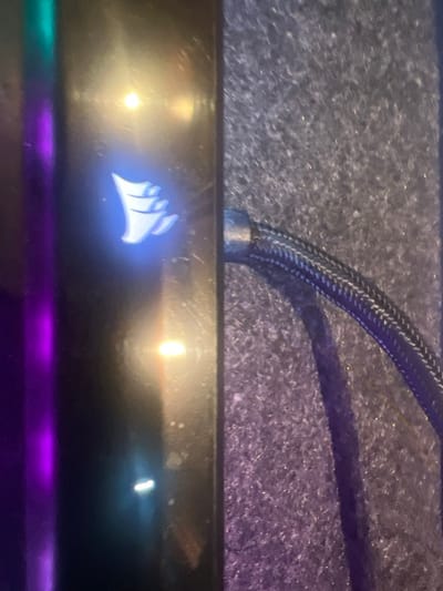 A close-up shows a Corsair computer component. The Corsair logo is illuminated in blue. Above, a braided cable runs across a textured, possibly carpeted, surface. Light reflections are visible on the component’s surface. A faint purple and teal glow emanates from below the component. The image appears to be taken in low light conditions.