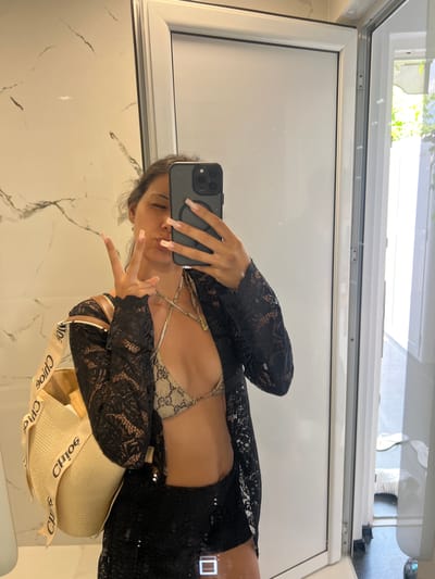 A woman is taking a mirror selfie in what appears to be a bathroom. She is wearing a black lace cover-up over a patterned bikini top and black bottoms. She is holding a straw bag with "Chloé" written on the strap. Her left hand is making a peace sign. The bathroom has marble-patterned walls and a white door. The location is not in the city of Chaidefto, Greece; it is a generic bathroom.