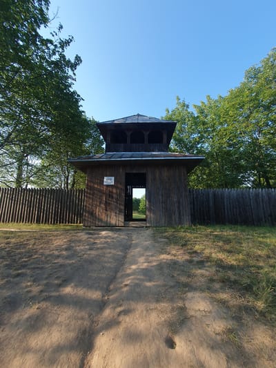 The image shows a wooden observation tower or structure, possibly part of a historical reconstruction or park, in Halych, Ukraine. The tower is dark wood, with a multi-tiered roof and open doorways. It is surrounded by trees with green foliage and a clear blue sky is visible. A wooden fence runs alongside the structure and continues into the background. The ground in front of the tower is a dirt path with some patches of dry grass. Shadows from trees are cast across the path, suggesting it is daytime, likely late afternoon or early morning due to the angle of the light. There is a small sign with text on the side of the tower, but it is too blurry to read. The scene appears peaceful and natural.